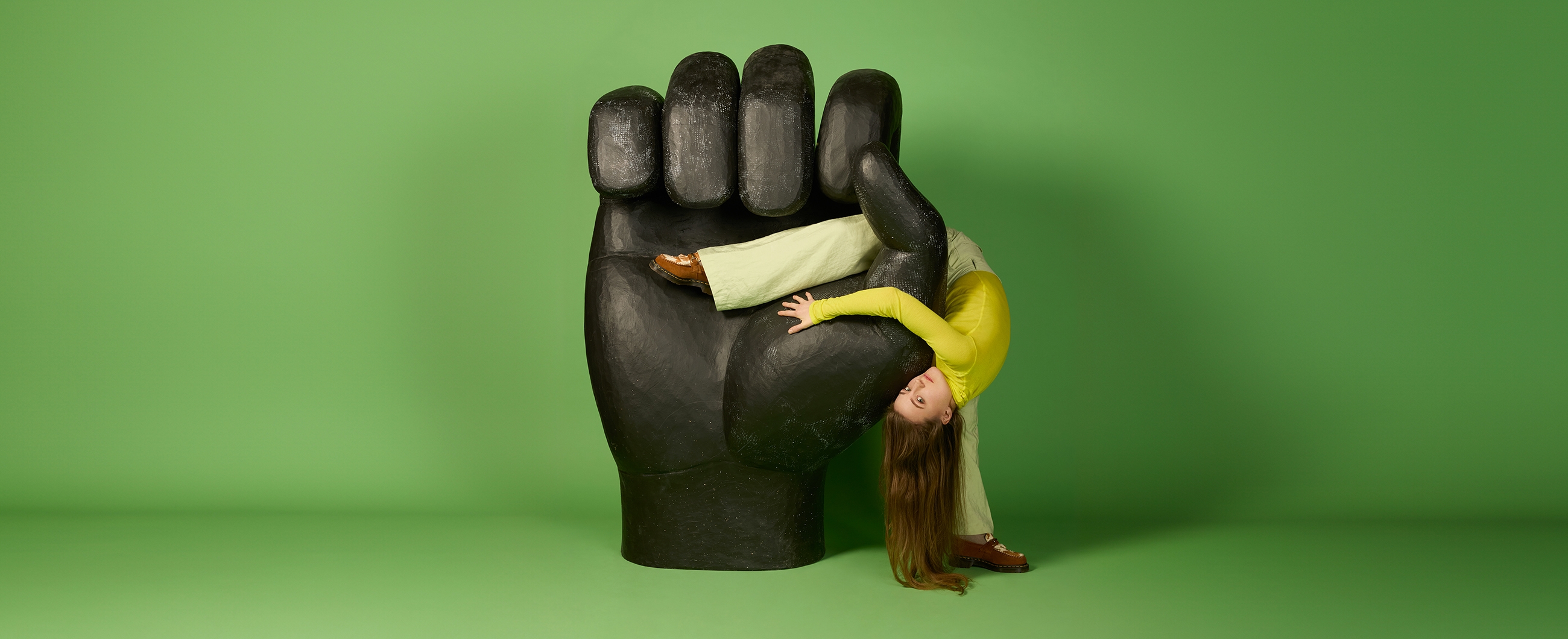 Eine Tänzerin schmiegt sich mit dem Kopf nach unten halb in eine riesige Hand-Skulptur. Sie blickt in die Kamera und ihr langes Haar reicht bis auf den Boden.